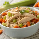 Shredded crockpot chicken with carrots and celery on wood.